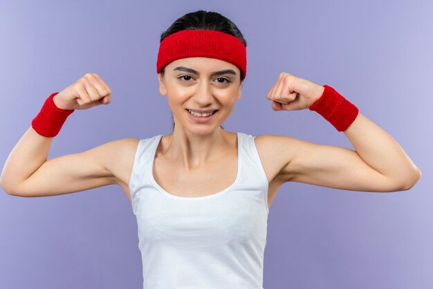 Mujer joven fitness en ropa deportiva con diadema sonriendo alegremente levantando puños mostrando bíceps feliz y positivo parado sobre pared púrpura