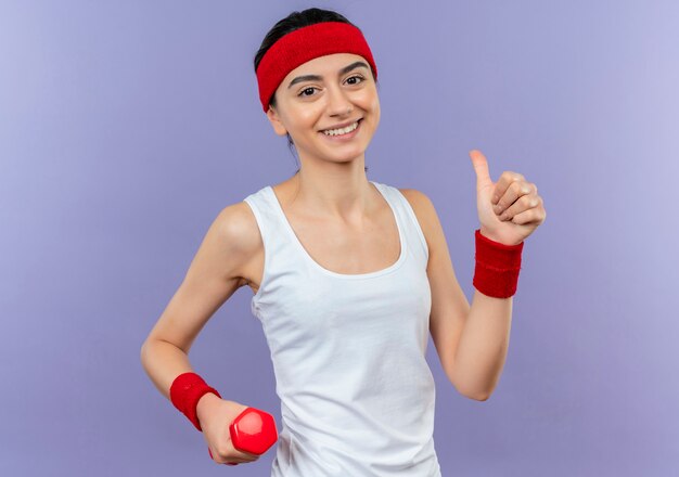 Mujer joven fitness en ropa deportiva con diadema sonriendo alegremente mostrando los pulgares para arriba de pie sobre la pared púrpura