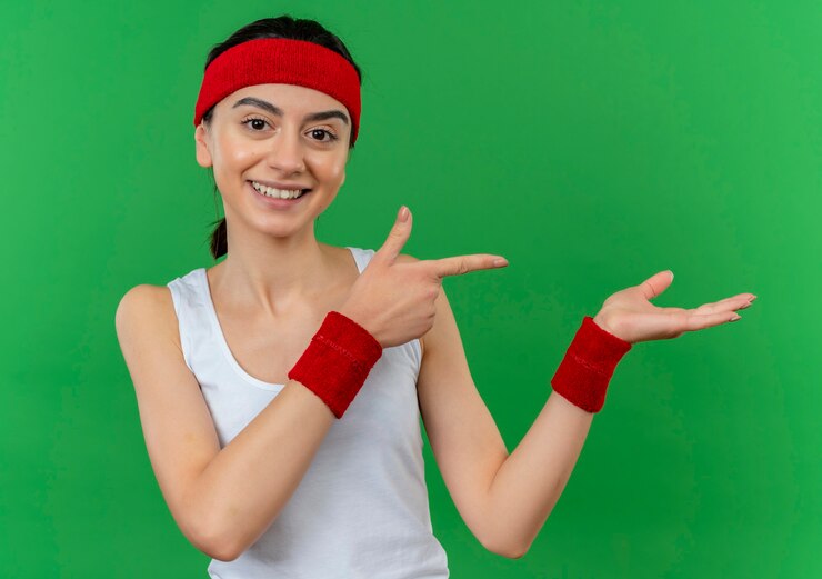 Foto gratuito mujer joven fitness en ropa deportiva con diadema sonriendo confiado apuntando con el dedo índice hacia el lado que presenta con el brazo de su mano de pie sobre la pared verde