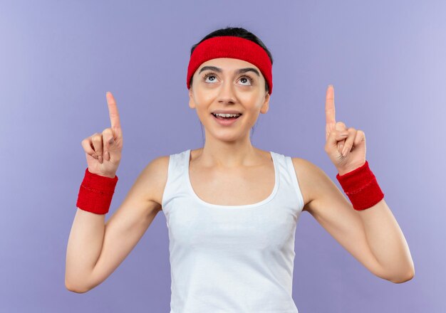 Mujer joven fitness en ropa deportiva con diadema sonriendo confiado apuntando con los dedos índices de pie sobre la pared púrpura