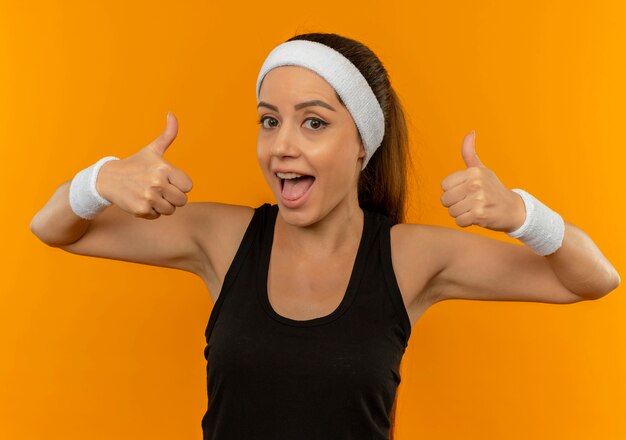 Mujer joven fitness en ropa deportiva con diadema sonriendo feliz y positivo mostrando los pulgares para arriba de pie sobre la pared naranja