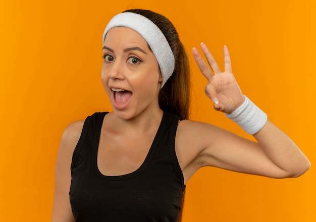 Mujer joven fitness en ropa deportiva con diadema sonriendo feliz y positivo mostrando signo ok de pie sobre la pared naranja