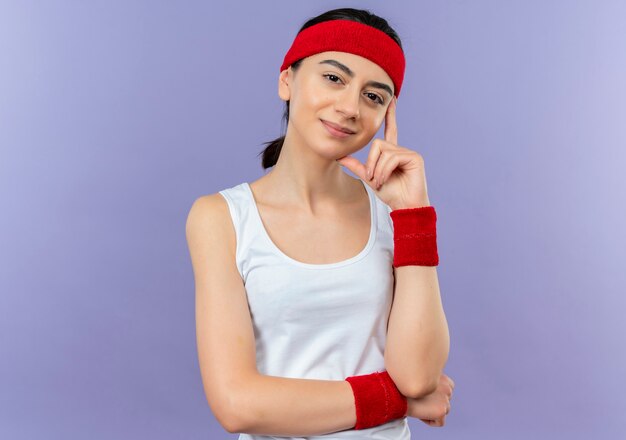 Mujer joven fitness en ropa deportiva con diadema con sonrisa de confianza en la cara de pie sobre la pared púrpura