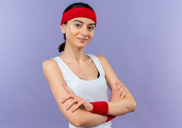 Mujer joven fitness en ropa deportiva con diadema con sonrisa de confianza con las manos cruzadas en el pecho de pie sobre la pared púrpura