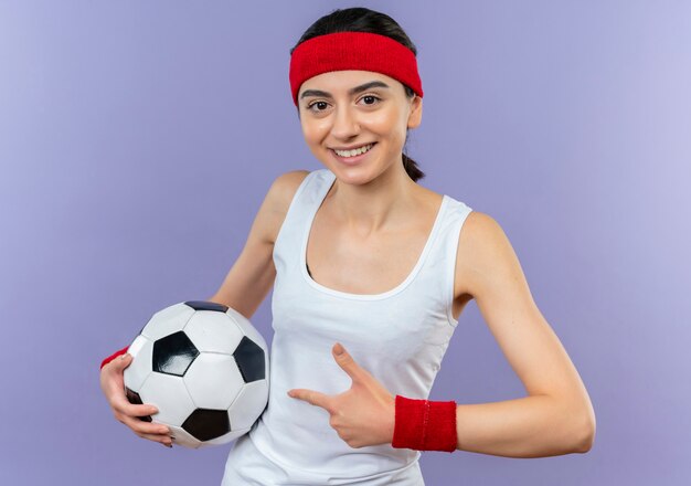 Mujer joven fitness en ropa deportiva con diadema sosteniendo un balón de fútbol apuntando con el dedo índice a él sonriendo de pie sobre la pared púrpura
