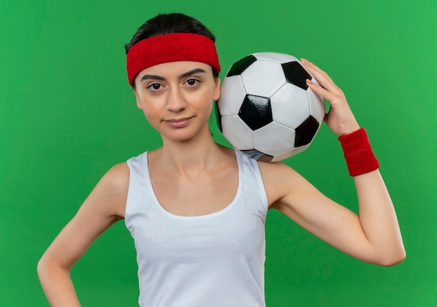 Mujer joven fitness en ropa deportiva con diadema sosteniendo un balón de fútbol con expresión de confianza de pie sobre la pared verde