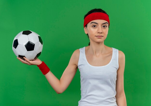 Mujer joven fitness en ropa deportiva con diadema sosteniendo un balón de fútbol con expresión de confianza de pie sobre la pared verde