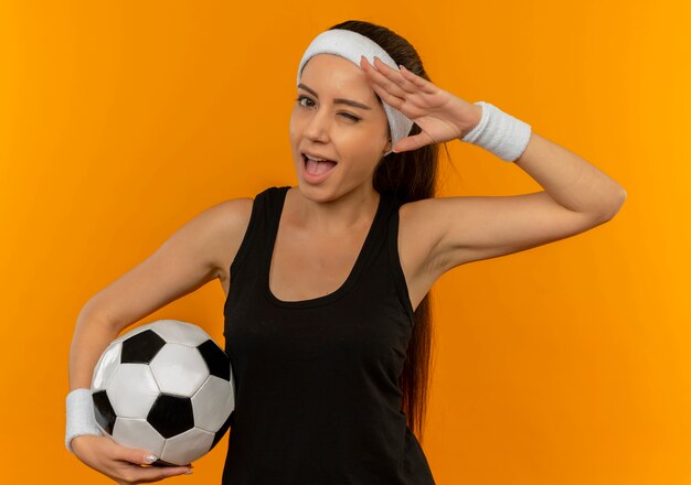 Mujer joven fitness en ropa deportiva con diadema sosteniendo un balón de fútbol guiñando un ojo y sonriendo, saludando de pie sobre la pared naranja