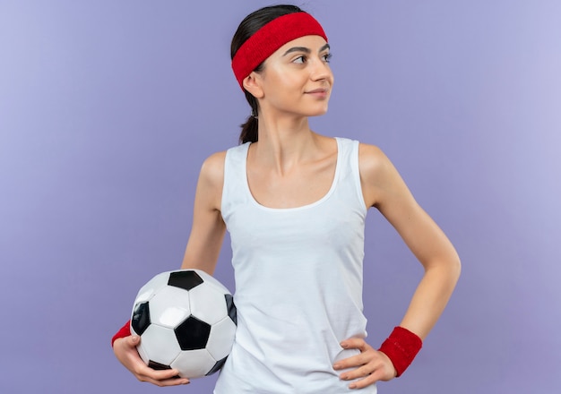 Mujer joven fitness en ropa deportiva con diadema sosteniendo un balón de fútbol mirando a un lado con sonrisa de confianza en la cara de pie sobre la pared púrpura