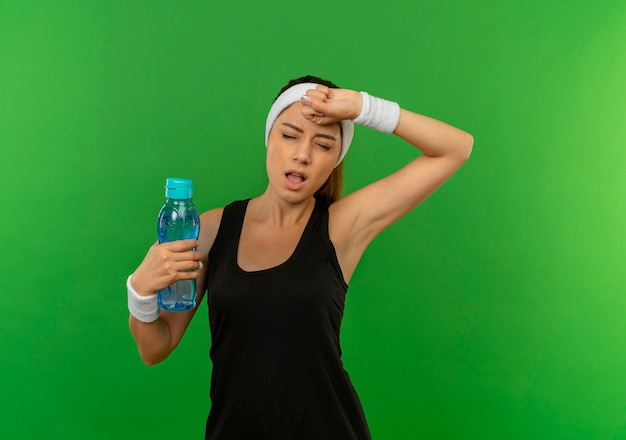 Mujer joven fitness en ropa deportiva con diadema sosteniendo una botella de agua con aspecto cansado y agotado de pie sobre la pared verde
