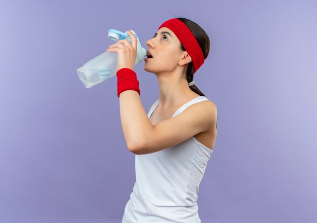 Mujer joven fitness en ropa deportiva con diadema sosteniendo una botella de agua con aspecto cansado de pie sobre la pared púrpura