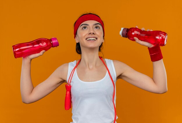 Mujer joven fitness en ropa deportiva con diadema sosteniendo dos botellas de agua mirando hacia arriba con una sonrisa en la cara de pie sobre la pared naranja