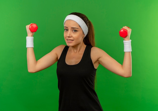 Mujer joven fitness en ropa deportiva con diadema sosteniendo dos mancuernas haciendo ejercicios mirando confundido parado sobre pared verde