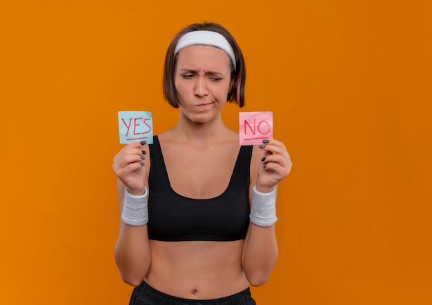 Mujer joven fitness en ropa deportiva con diadema sosteniendo dos papeles recordatorios con la palabra sí y no en manos levantadas mirándolos con expresión escéptica de pie sobre la pared naranja