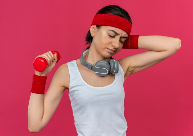 Mujer joven fitness en ropa deportiva con diadema sosteniendo mancuernas haciendo ejercicios mirando confundido malestar de pie sobre la pared rosa