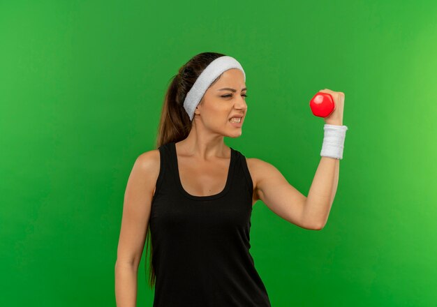 Mujer joven fitness en ropa deportiva con diadema sosteniendo mancuernas haciendo ejercicios mirando tenso parado sobre pared verde