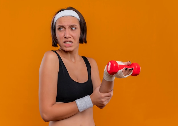 Mujer joven fitness en ropa deportiva con diadema sosteniendo mancuernas tocando su muñeca con aspecto enfermo y dolor de pie sobre la pared naranja