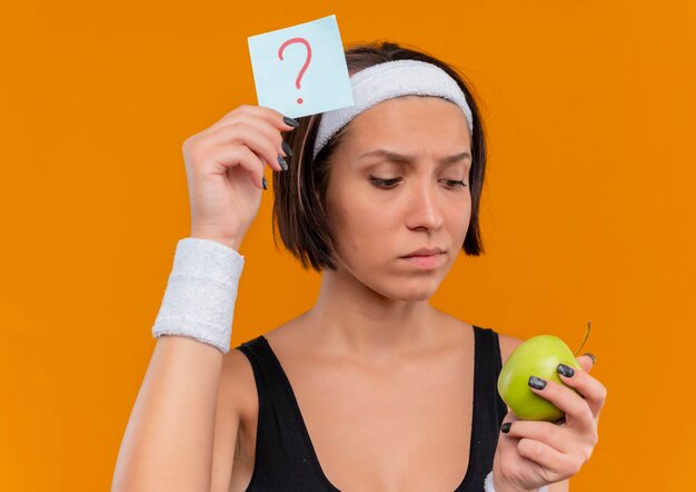 Mujer joven fitness en ropa deportiva con diadema sosteniendo papel recordatorio con signo de interrogación y manzana verde mirando manzana con expresión pensativa de pie sobre la pared naranja