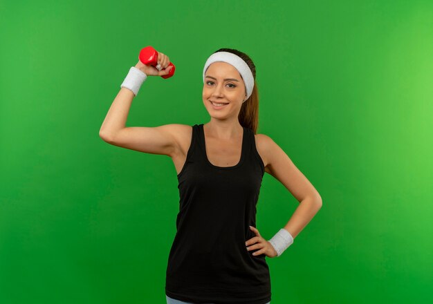 Mujer joven fitness en ropa deportiva con diadema sosteniendo pesas haciendo ejercicios mirando confiado con una sonrisa en la cara de pie sobre la pared verde