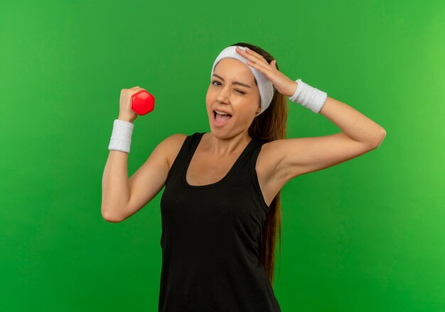 Mujer joven fitness en ropa deportiva con diadema sosteniendo pesas haciendo ejercicios sonriendo y guiñando un ojo de pie sobre la pared verde