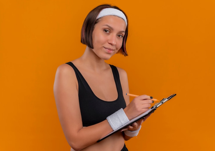 Foto gratuito mujer joven fitness en ropa deportiva con diadema sosteniendo portapapeles con páginas en blanco escribiendo mirando confiado de pie sobre la pared naranja