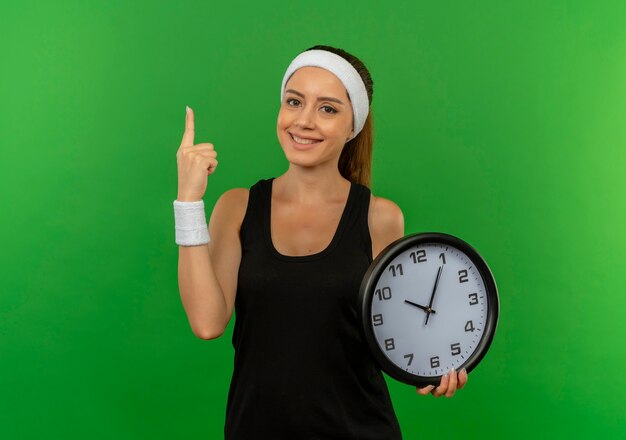 Mujer joven fitness en ropa deportiva con diadema sosteniendo reloj de pared mostrando el dedo índice sonriendo confiado de pie sobre la pared verde