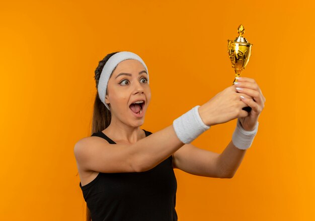 Mujer joven fitness en ropa deportiva con diadema sosteniendo trofeo feliz y emocionado de pie sobre la pared naranja