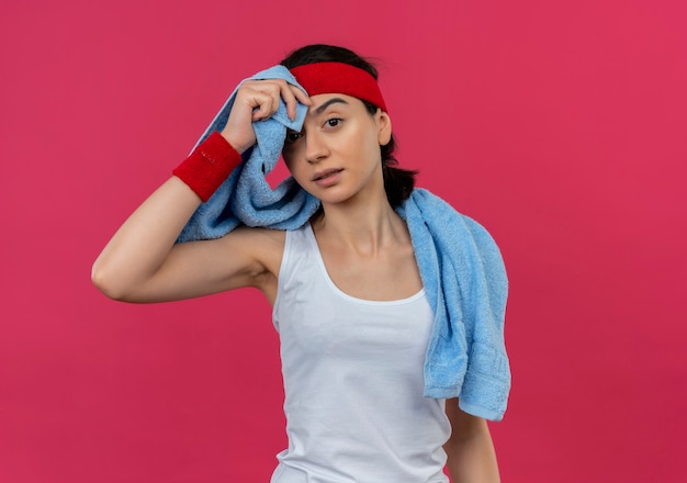 Mujer joven fitness en ropa deportiva con diadema y toalla alrededor de su cuello cansado y agotado de pie sobre la pared rosa