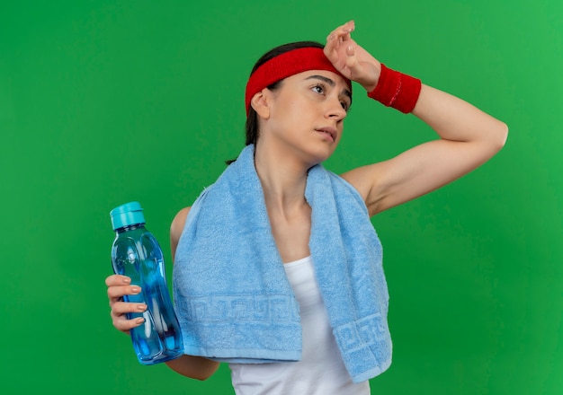 Mujer joven fitness en ropa deportiva con diadema y toalla en el cuello sosteniendo una botella de agua con aspecto cansado y agotado de pie sobre la pared verde