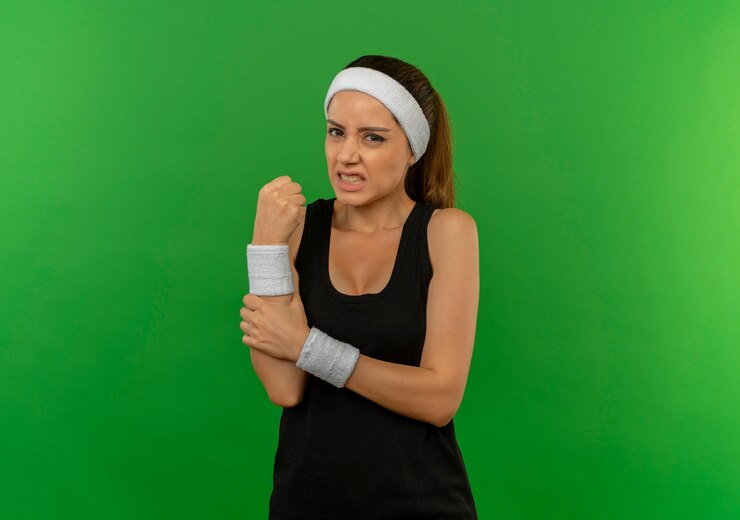 Foto gratuito mujer joven fitness en ropa deportiva con diadema tocando su muñeca con dolor de pie sobre la pared verde