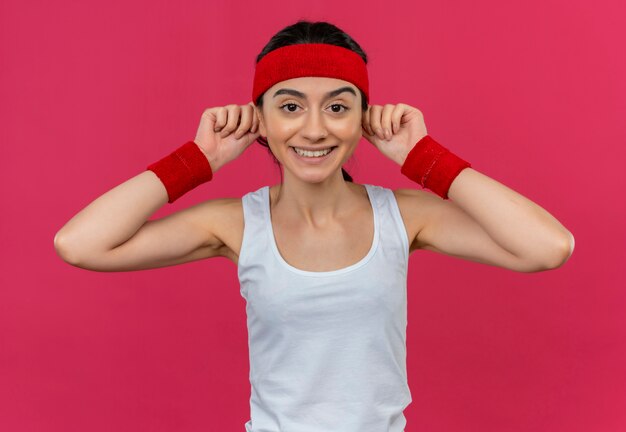 Mujer joven fitness en ropa deportiva con diadema tocando sus orejas con una gran sonrisa en la cara de pie sobre la pared rosa