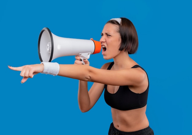 Mujer joven fitness en ropa deportiva gritando al megáfono con expresión agresiva apuntando con el dedo índice a algo parado sobre la pared azul