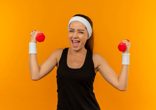 Mujer joven fitness en ropa deportiva haciendo ejercicios con dos pesas sonriendo alegremente feliz y emocionado de pie sobre la pared naranja