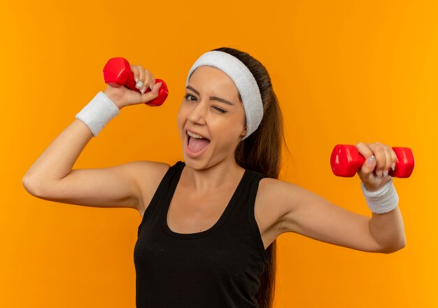 Mujer joven fitness en ropa deportiva haciendo ejercicios con dos pesas sonriendo alegremente de pie sobre la pared naranja