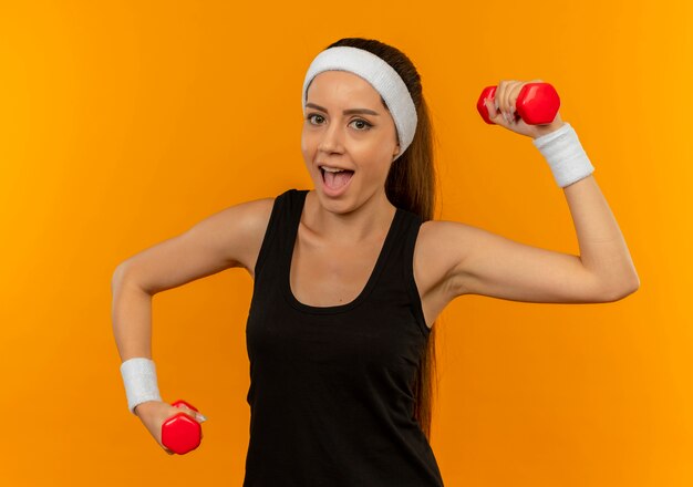 Mujer joven fitness en ropa deportiva haciendo ejercicios con dos pesas sonriendo alegremente de pie sobre la pared naranja