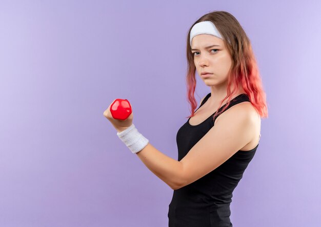 Mujer joven fitness en ropa deportiva haciendo ejercicios con mancuernas con rostro serio de pie sobre la pared púrpura