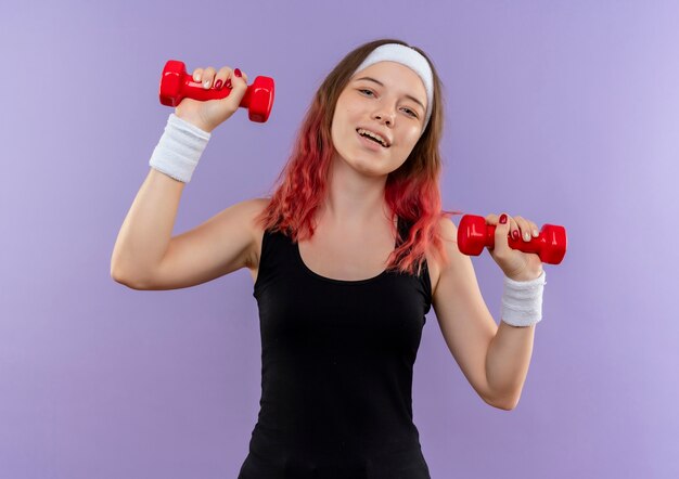 Mujer joven fitness en ropa deportiva haciendo ejercicios con mancuernas sonriendo alegremente de pie sobre la pared púrpura