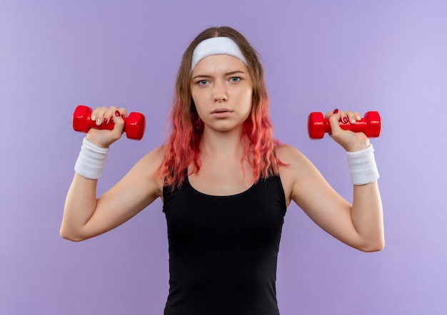 Foto gratuita mujer joven fitness en ropa deportiva haciendo ejercicios con pesas con rostro serio de pie sobre la pared púrpura