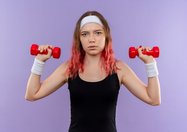 Mujer joven fitness en ropa deportiva haciendo ejercicios con pesas con rostro serio de pie sobre la pared púrpura