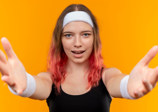 Foto gratuita mujer joven fitness en ropa deportiva haciendo gesto de bienvenida con las manos de pie sobre la pared naranja
