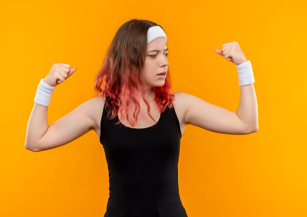 Mujer joven fitness en ropa deportiva levantando puños mostrando bíceps mirando confiado parado sobre pared naranja