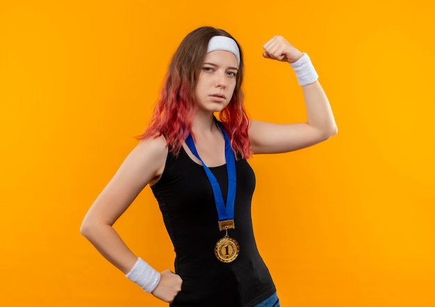 Mujer joven fitness en ropa deportiva con medalla de oro alrededor de su cuello levantando el puño mirando confiado mostrando bíceps