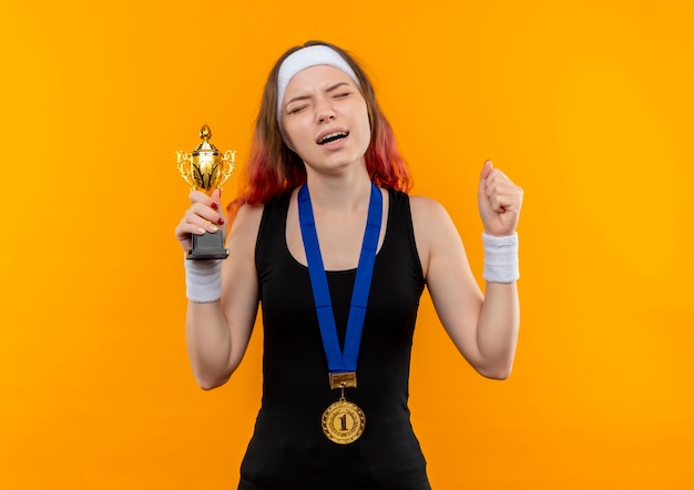 Foto gratuita mujer joven fitness en ropa deportiva con medalla de oro alrededor de su cuello levantando puños sosteniendo el trofeo con expresión molesta de pie sobre la pared naranja