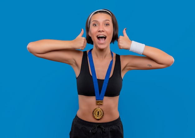 Foto gratuita mujer joven fitness en ropa deportiva con medalla de oro alrededor de su cuello mostrando los pulgares para arriba regocijándose de su éxito feliz y emocionada de pie sobre la pared azul