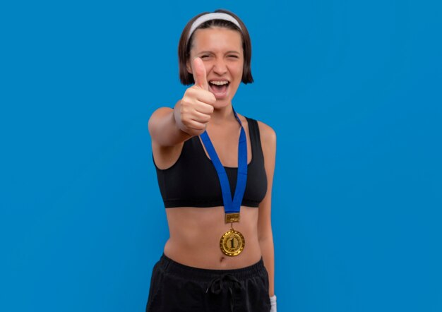 Mujer joven fitness en ropa deportiva con medalla de oro alrededor de su cuello mostrando los pulgares para arriba regocijándose de su éxito feliz y emocionada de pie sobre la pared azul