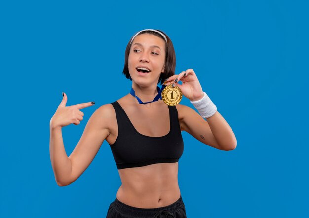 Mujer joven fitness en ropa deportiva con medalla de oro alrededor de su cuello que muestra la medalla apuntando con el dedo índice a ella sonriendo confiada con orgullo de pie sobre la pared azul