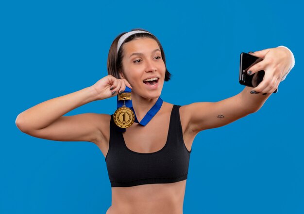 Mujer joven fitness en ropa deportiva con medalla de oro alrededor de su cuello tomando selfie mostrando medalla a la cámara de su teléfono inteligente de pie sobre la pared azul