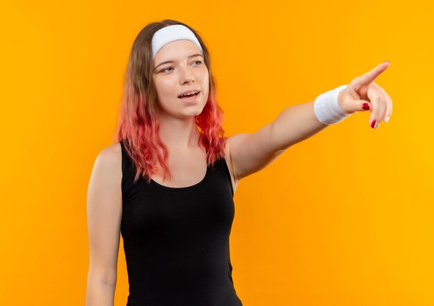 Mujer joven fitness en ropa deportiva mirando a un lado apuntando con el dedo a algo sonriente de pie sobre la pared naranja