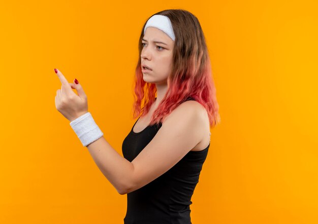 Mujer joven fitness en ropa deportiva mirando a un lado apuntando con el dedo con expresión confusa de pie sobre la pared naranja