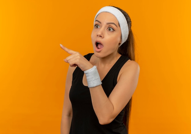 Mujer joven fitness en ropa deportiva mirando sorprendido apuntando con el dedo hacia el lado parado sobre la pared naranja
